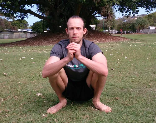 A man resting in a deep squat on the grass in a park